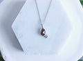 A silver necklace with a teardrop-shaped brown gemstone pendant and a small clear accent stone, displayed on a white hexagonal stand with blurred green foliage in the background.