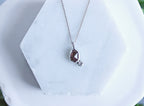 A silver necklace with a teardrop-shaped brown gemstone pendant and a small clear accent stone, displayed on a white hexagonal stand with blurred green foliage in the background.