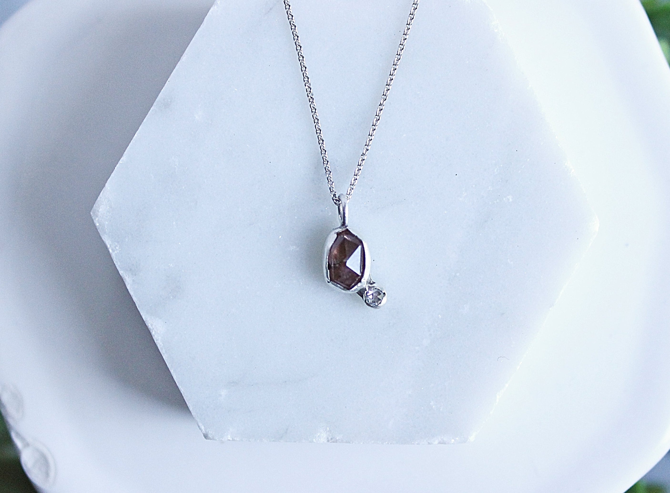 A silver necklace with a teardrop-shaped brown gemstone pendant and a small clear accent stone, displayed on a white hexagonal stand with blurred green foliage in the background.